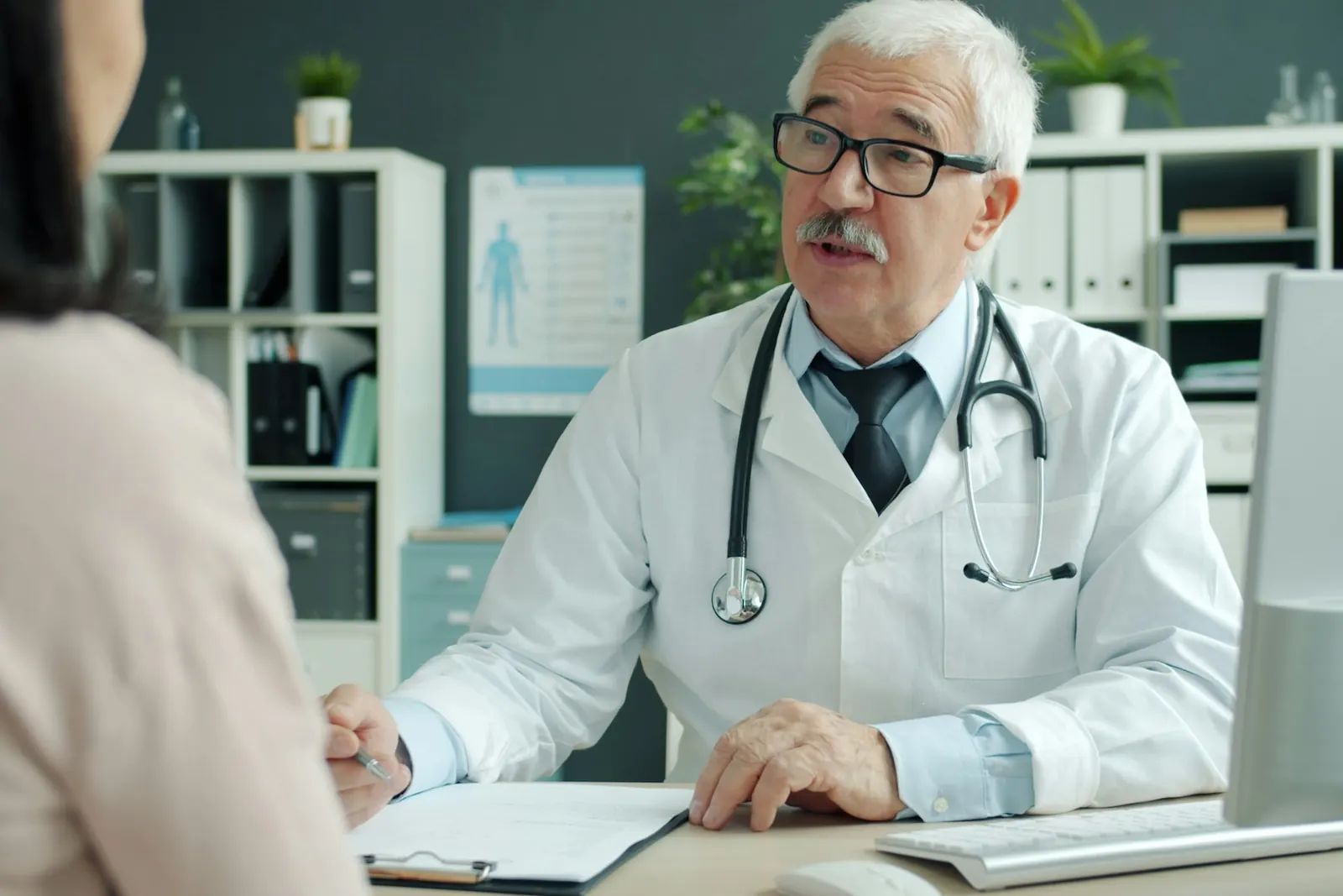 A doctor in a white coat talking at his desk with the person who has come for a consultation